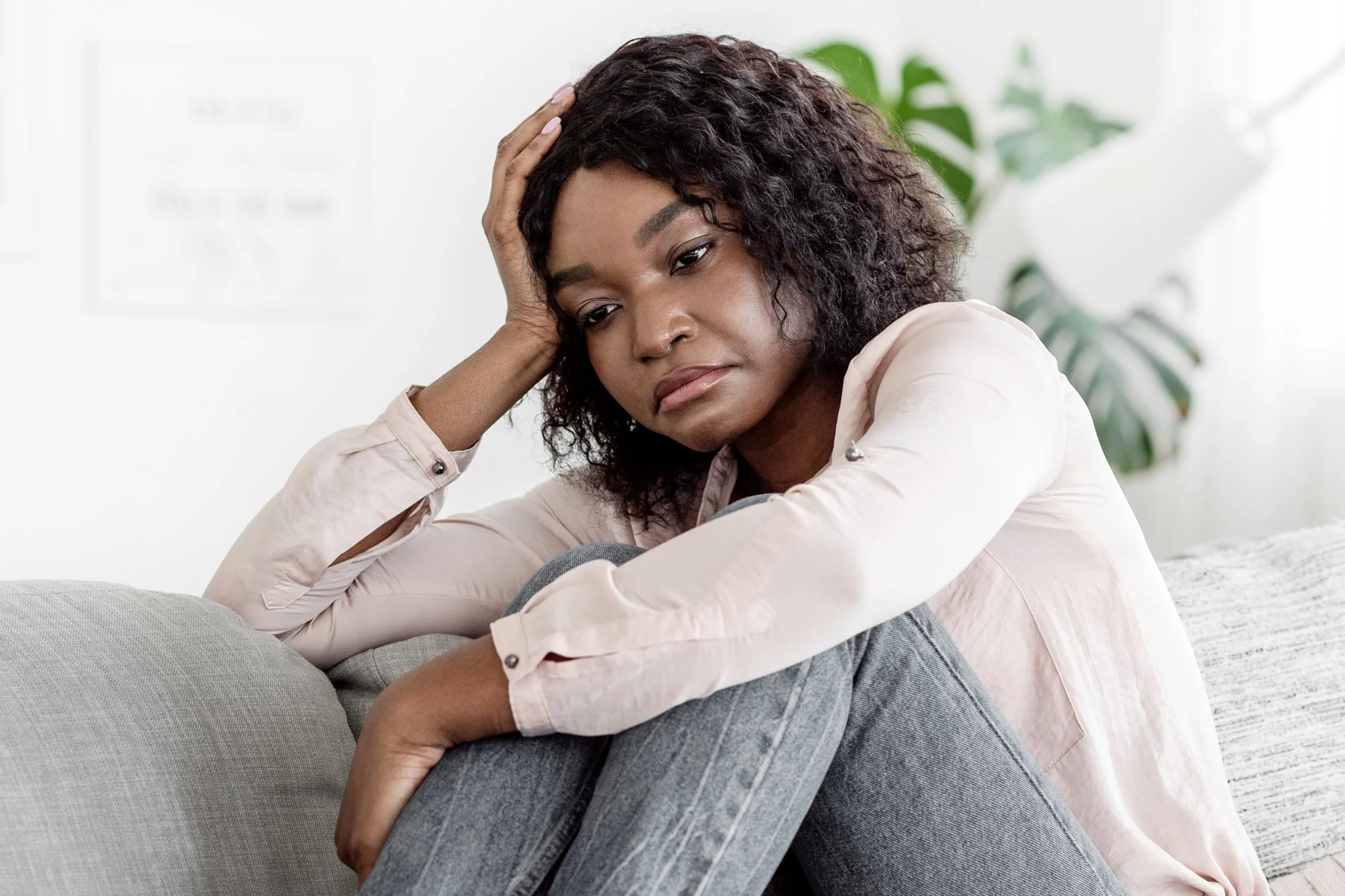 Woman in light pink shirt sitting on a bed with head in hand, looking downward with concerned expression