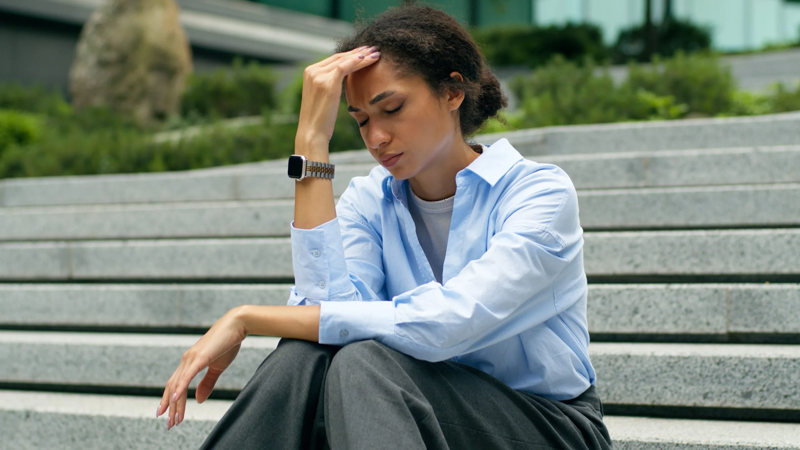 Woman sitting on outdoor steps with hand to forehead, appearing stressed or fatigued