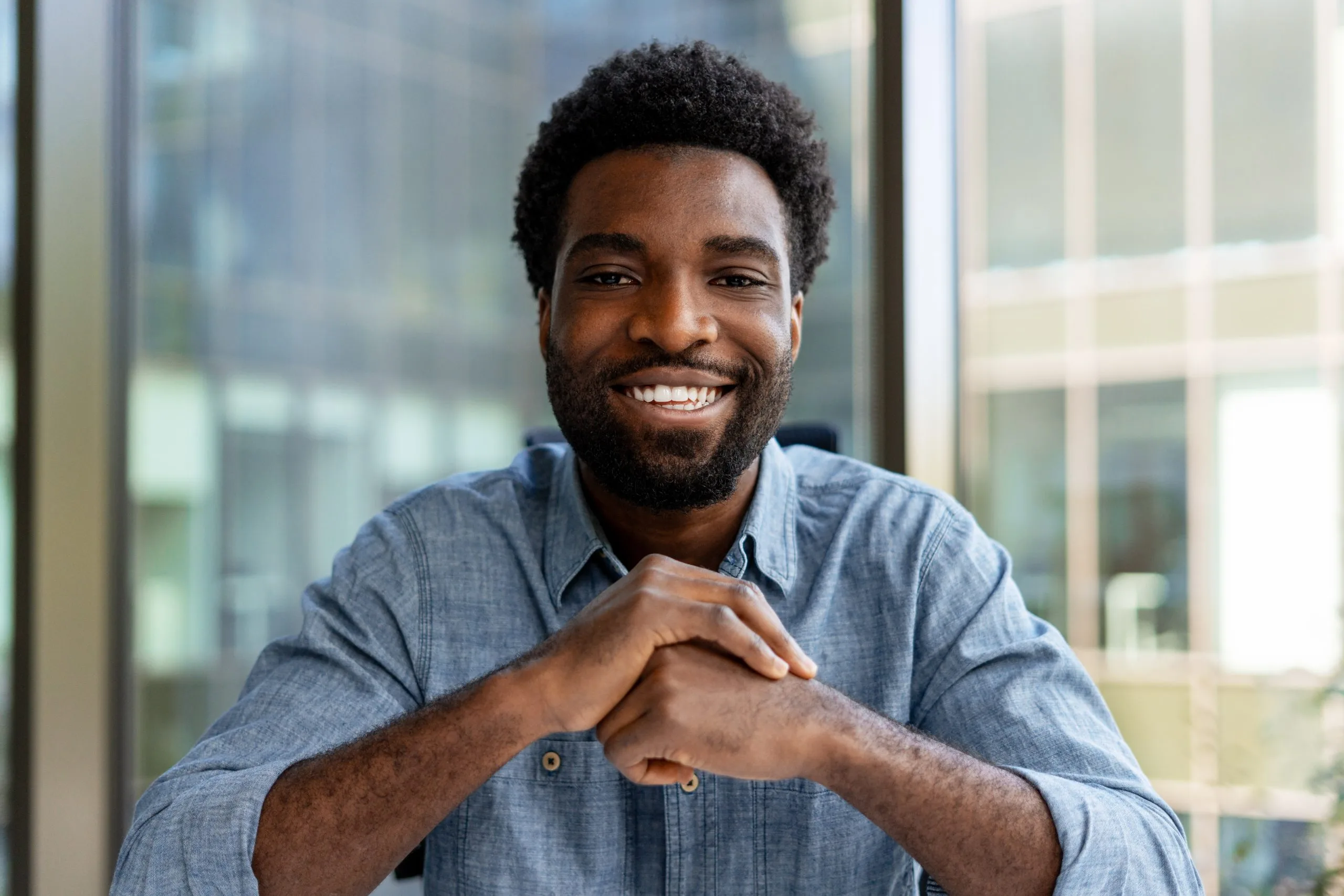 Man in blue shirt smiling at camera with hands clasped, standing by office windows