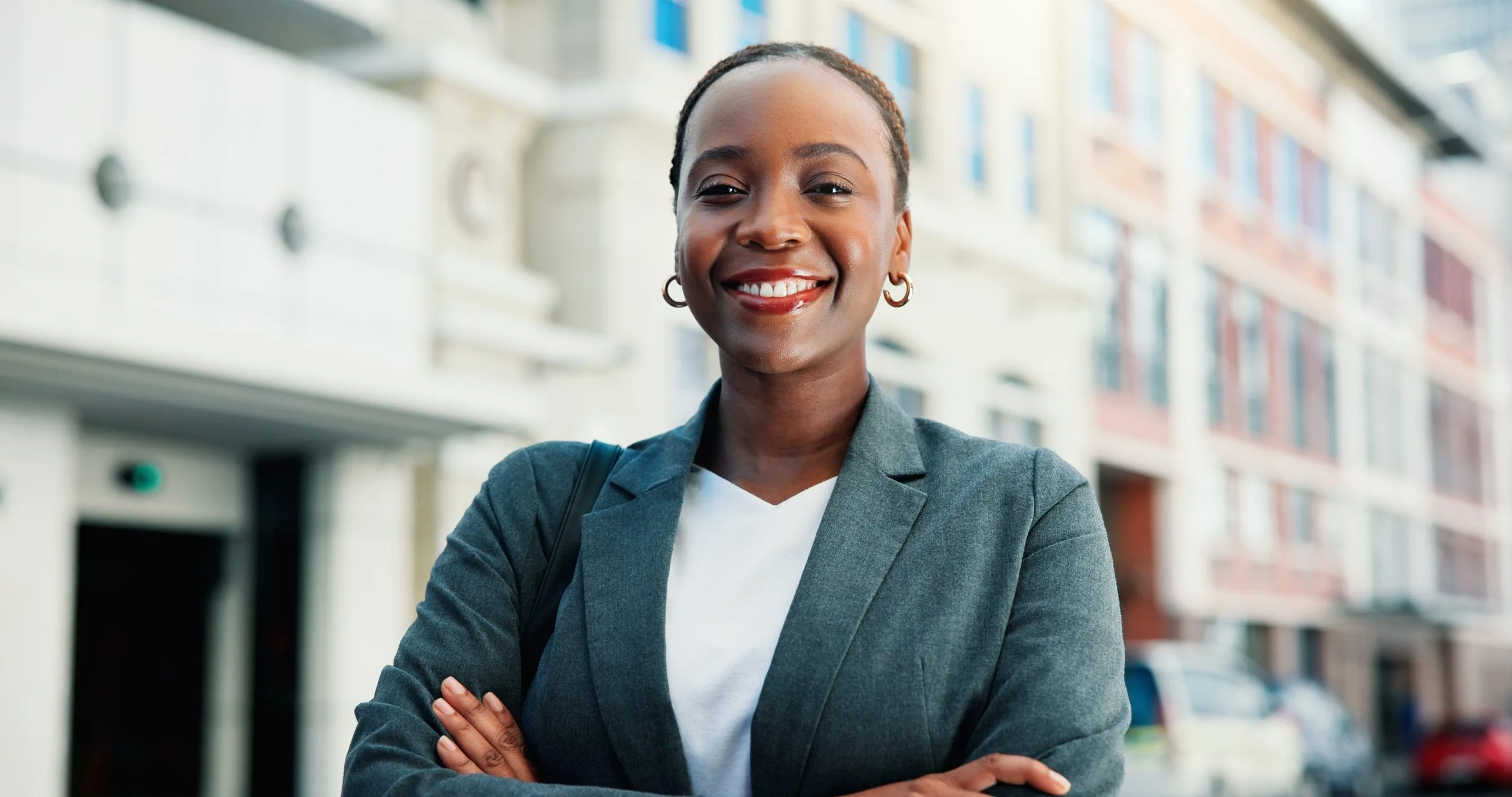 Smiling woman in teal blazer and white shirt with arms crossed standing outdoors in urban setting