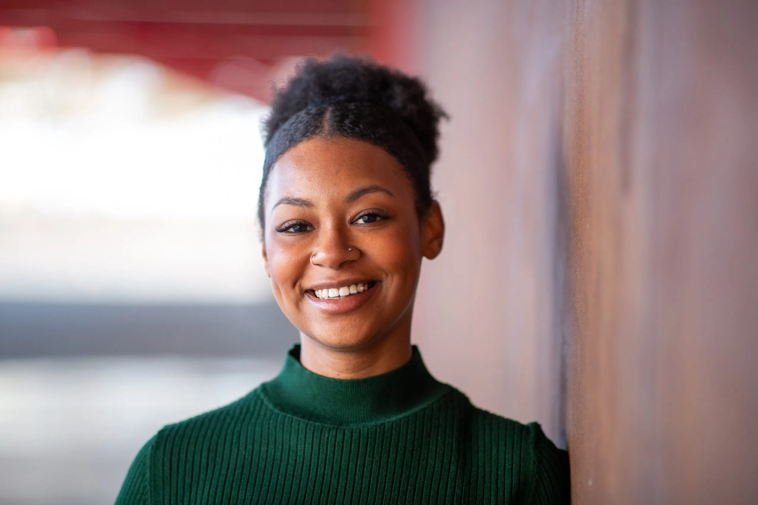 Smiling woman in dark green turtleneck standing indoors against pink wall