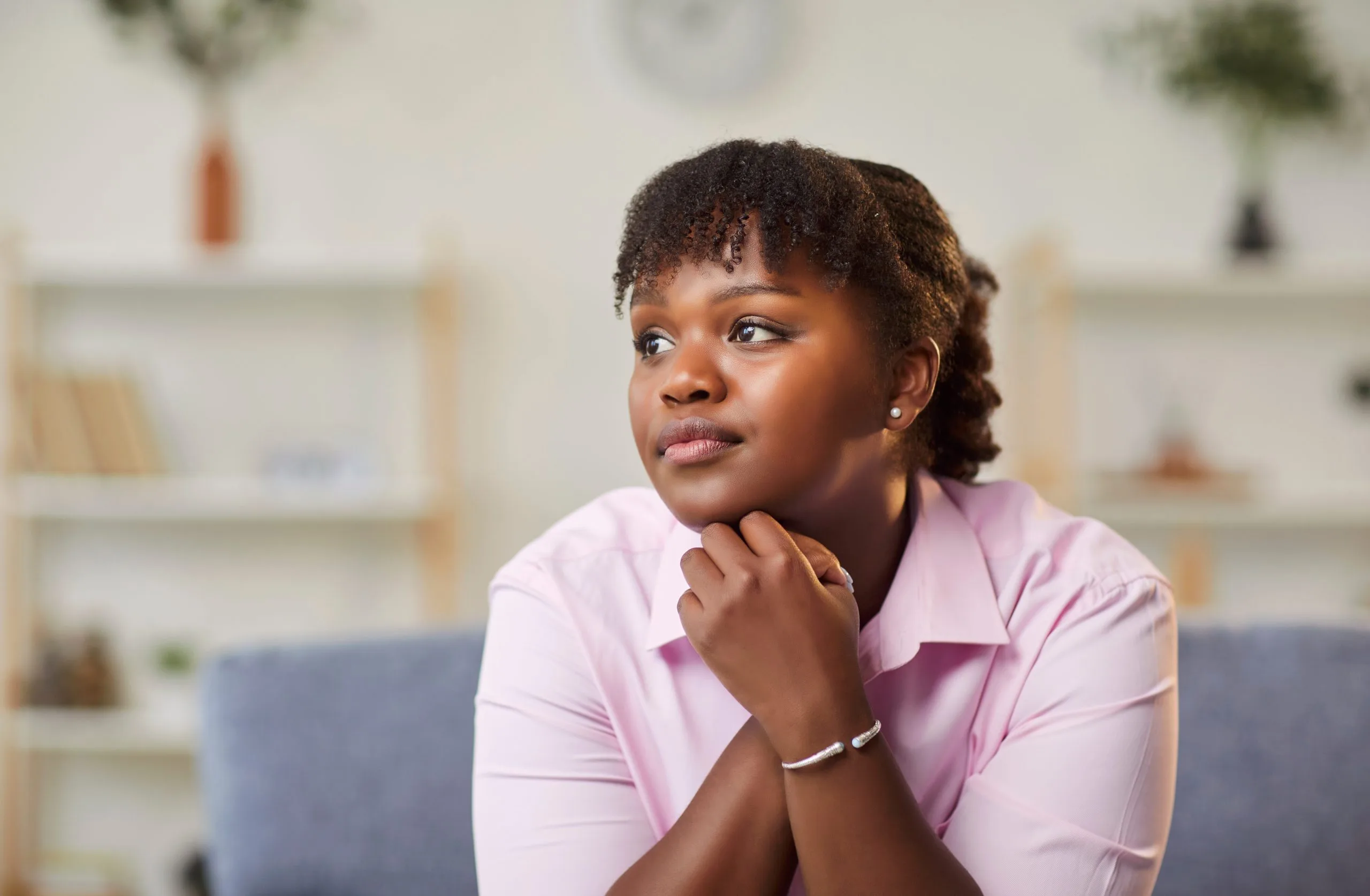 Woman in pink shirt sitting on couch with hand to chin, contemplative expression in modern living room