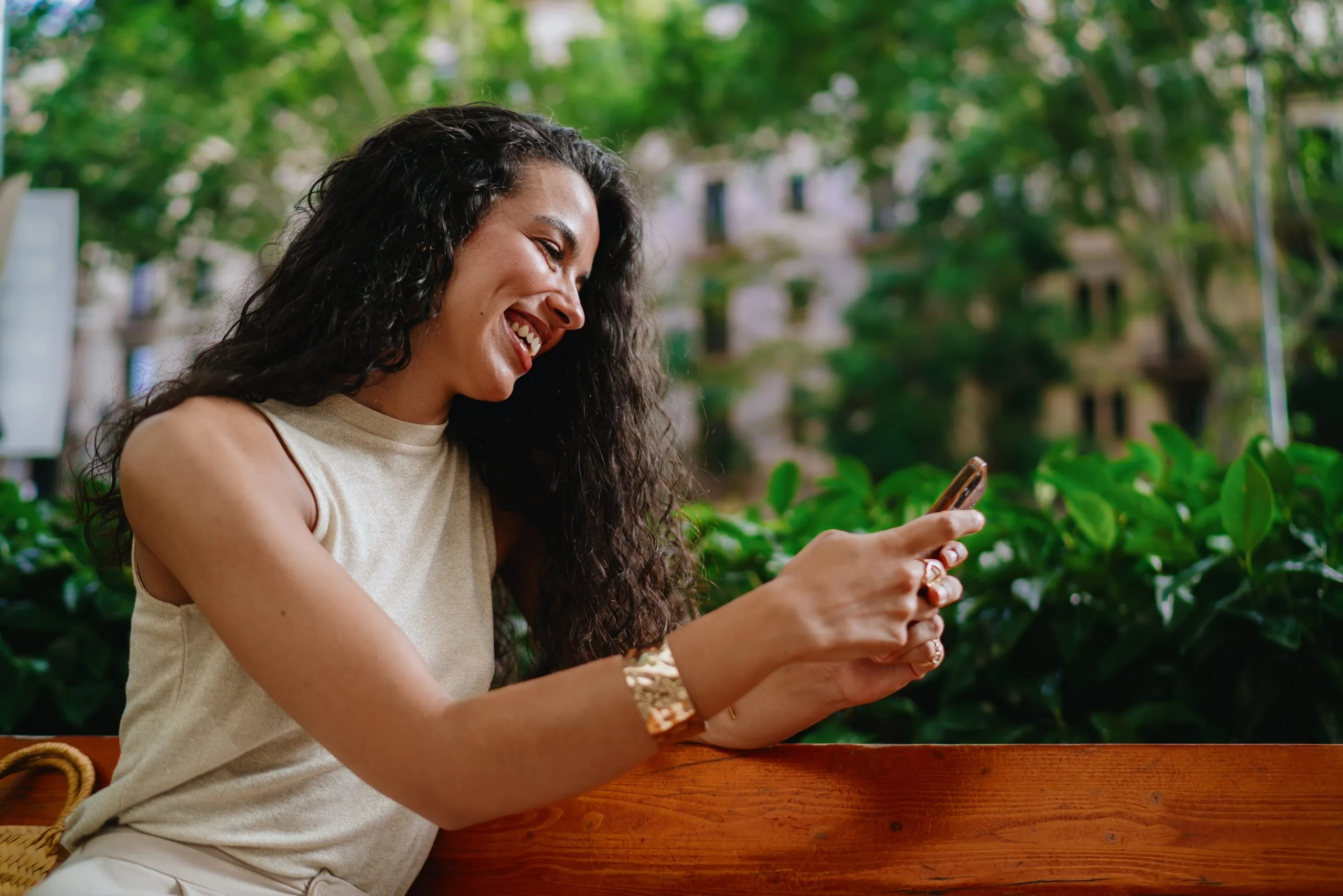 Woman with long dark hair smiling while using smartphone, seated outdoors with urban garden backdrop