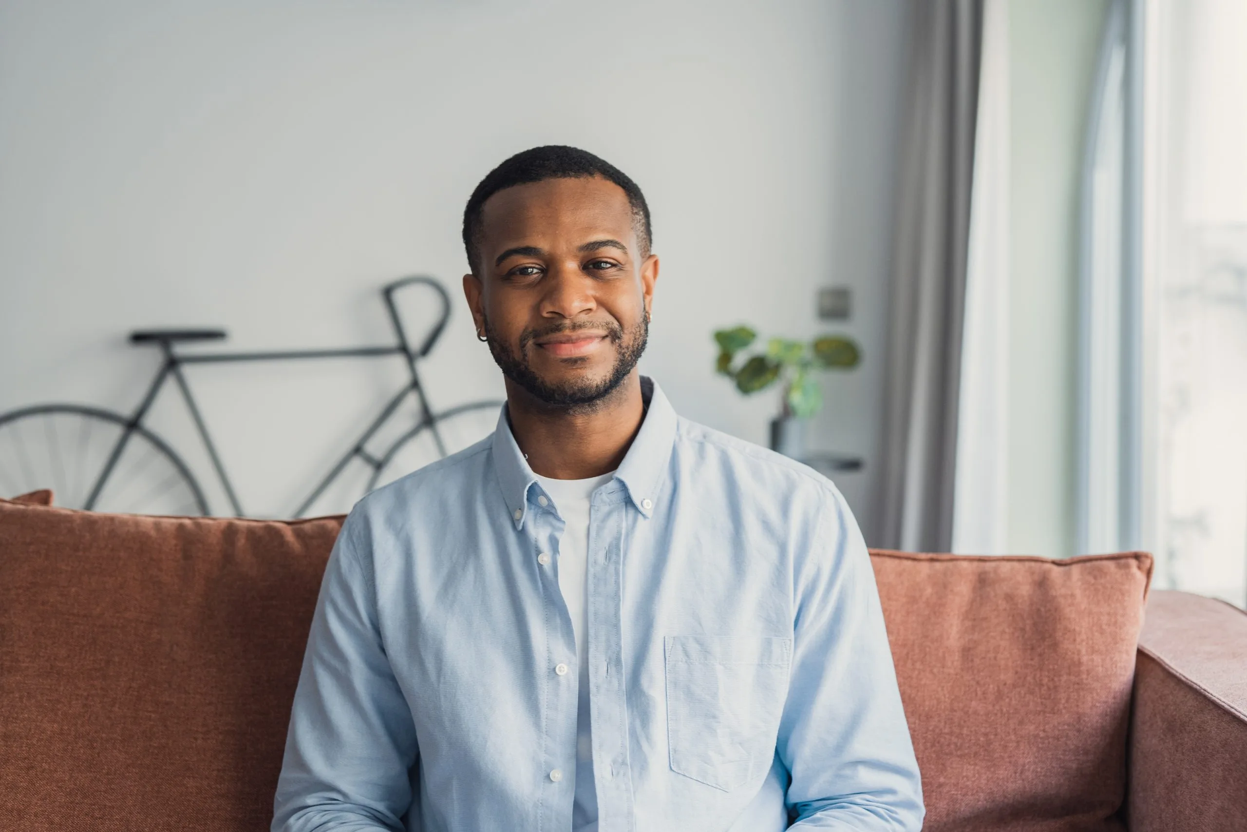 Man in light blue button-up shirt smiling at camera seated on rust-colored couch in minimalist home