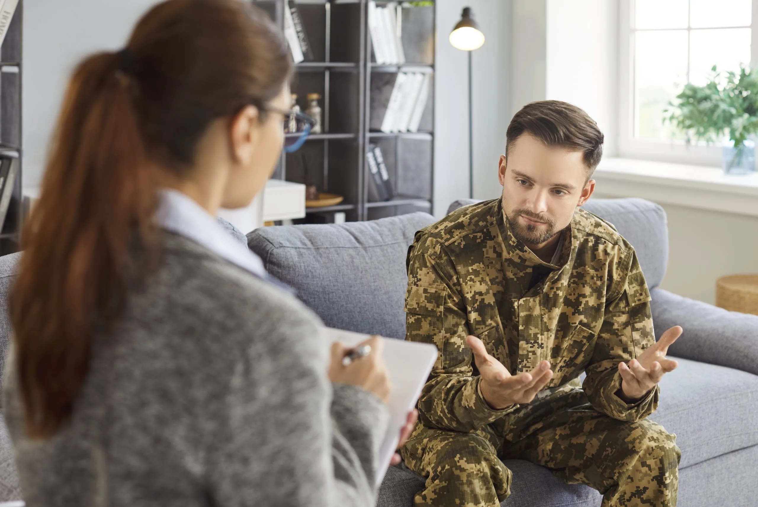 Female clinician with clipboard speaking with male service member in patterned shirt during office consultation