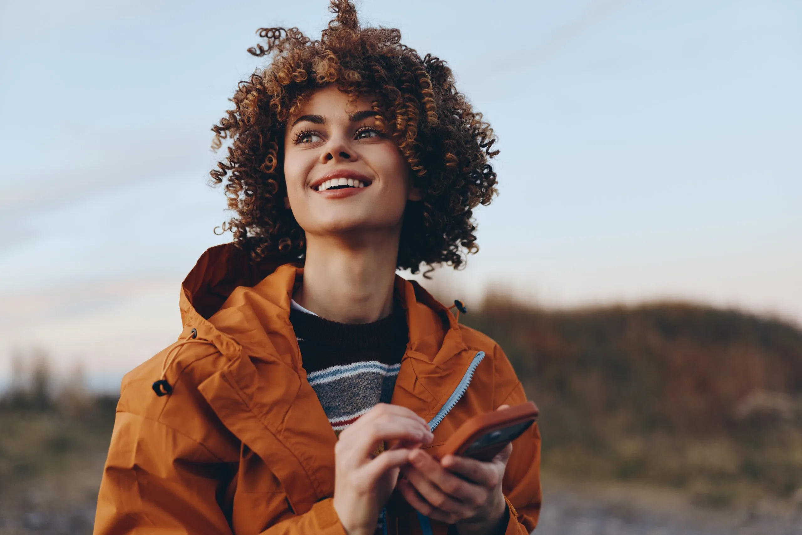 Woman with curly hair in orange jacket holding smartphone, smiling outdoors with forested hills behind