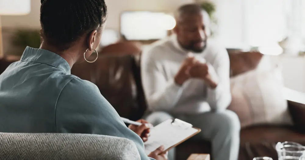 Therapist taking notes on clipboard during counseling session with two clients in an office setting
