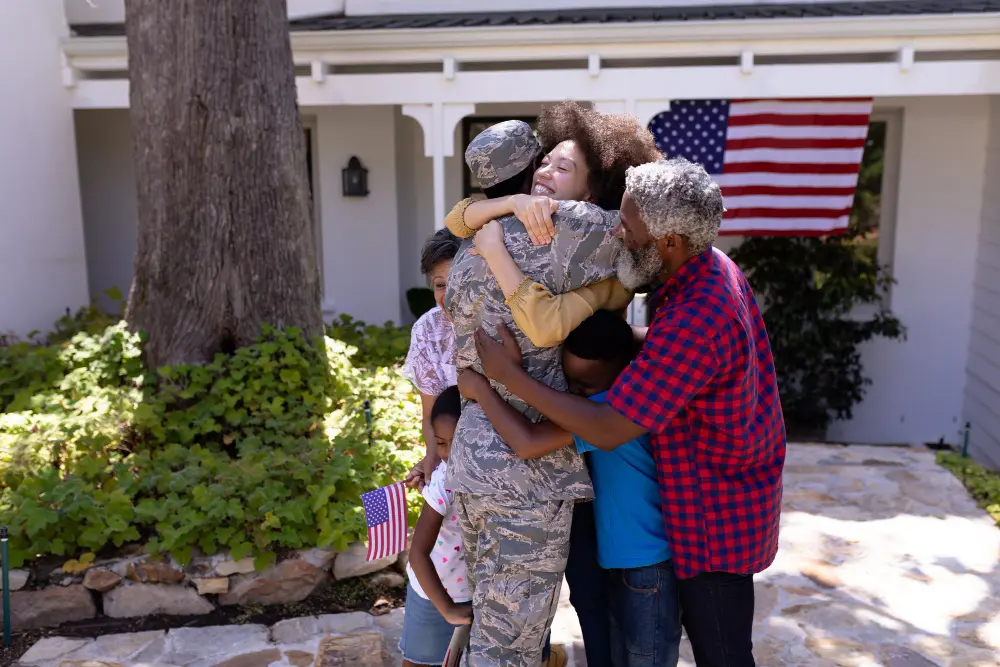 Military service member in uniform embracing family members outside home with American flag displayed