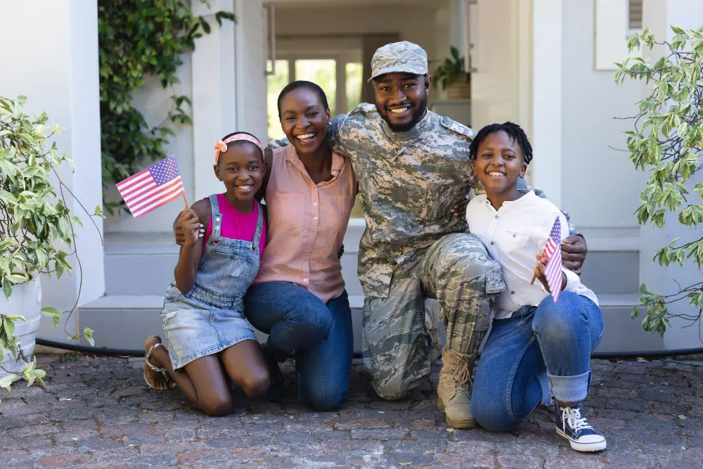 Military service member kneeling with three family members holding American flags outside residential home
