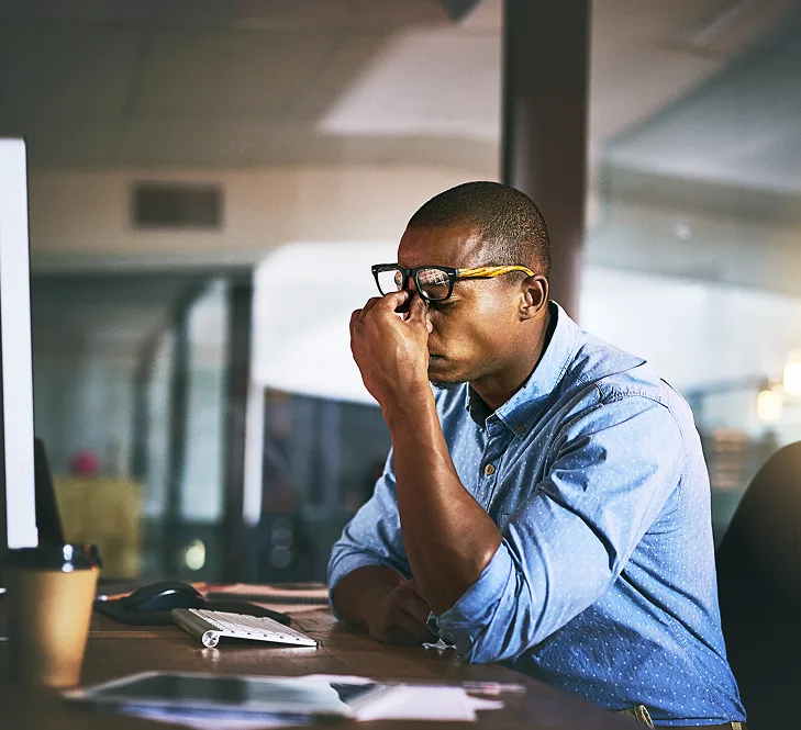Man in glasses at desk with computer, hand to face in thoughtful pose