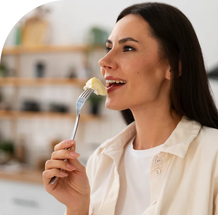 Woman eating food on a fork in a bright kitchen with wooden shelving in the background