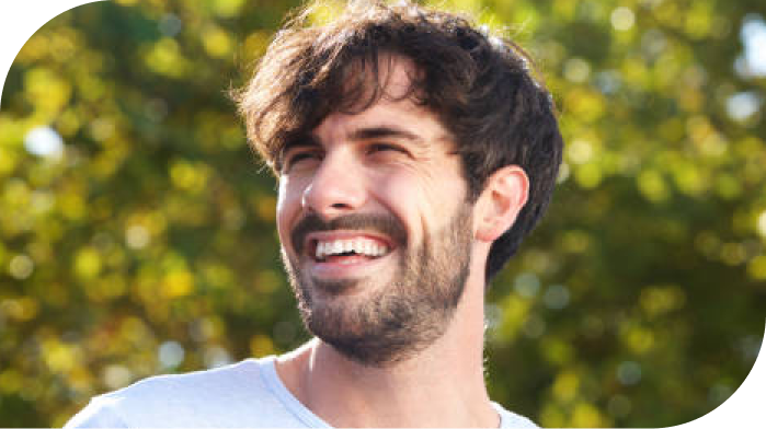 Smiling man with dark hair and beard outdoors with blurred green foliage background