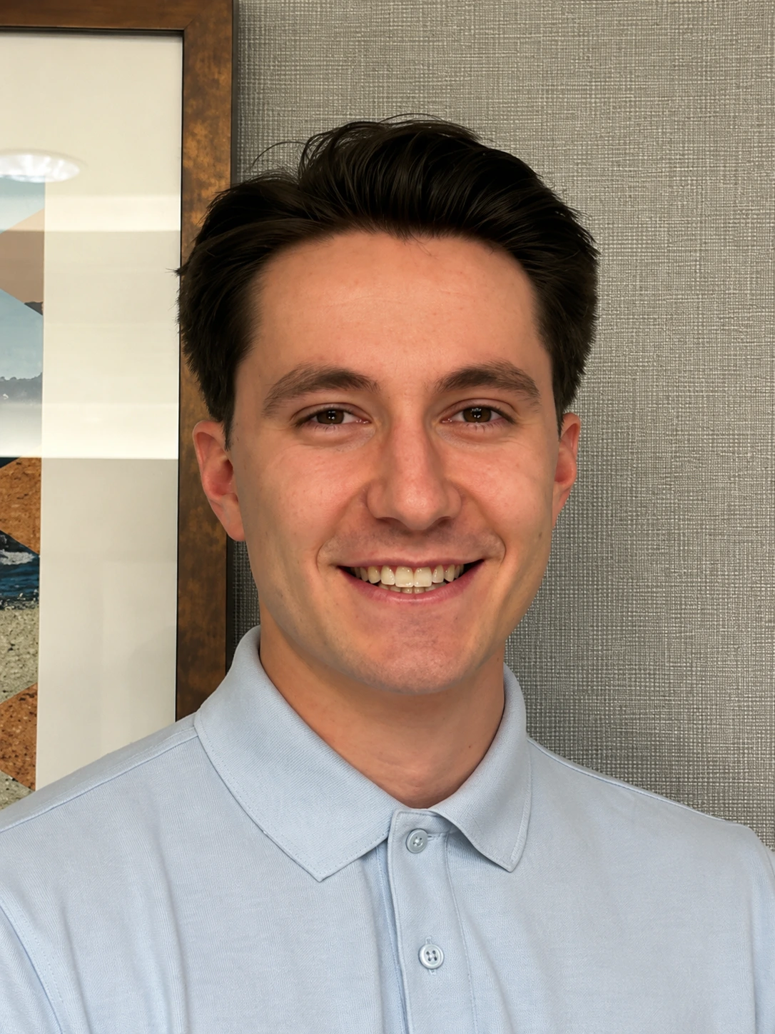 Man in light blue polo shirt smiling at camera in office with framed artwork on wall