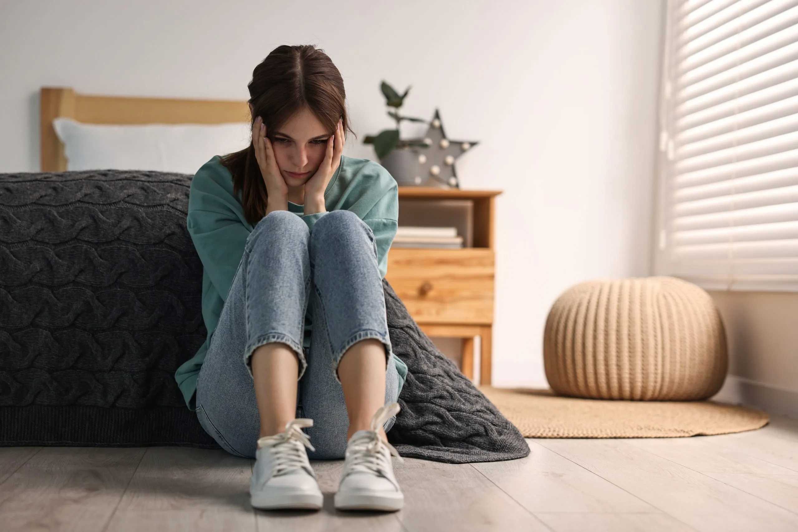 Teen girl sitting on bedroom floor with head in hands, appearing distressed
