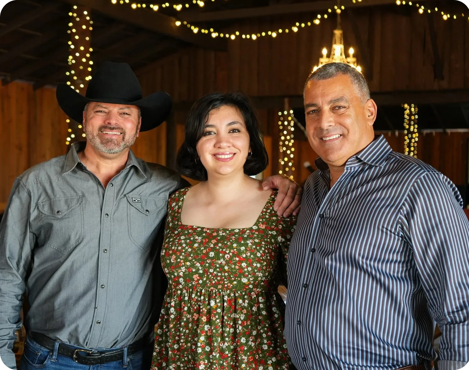 Three people smiling at camera in front of holiday-decorated barn with string lights
