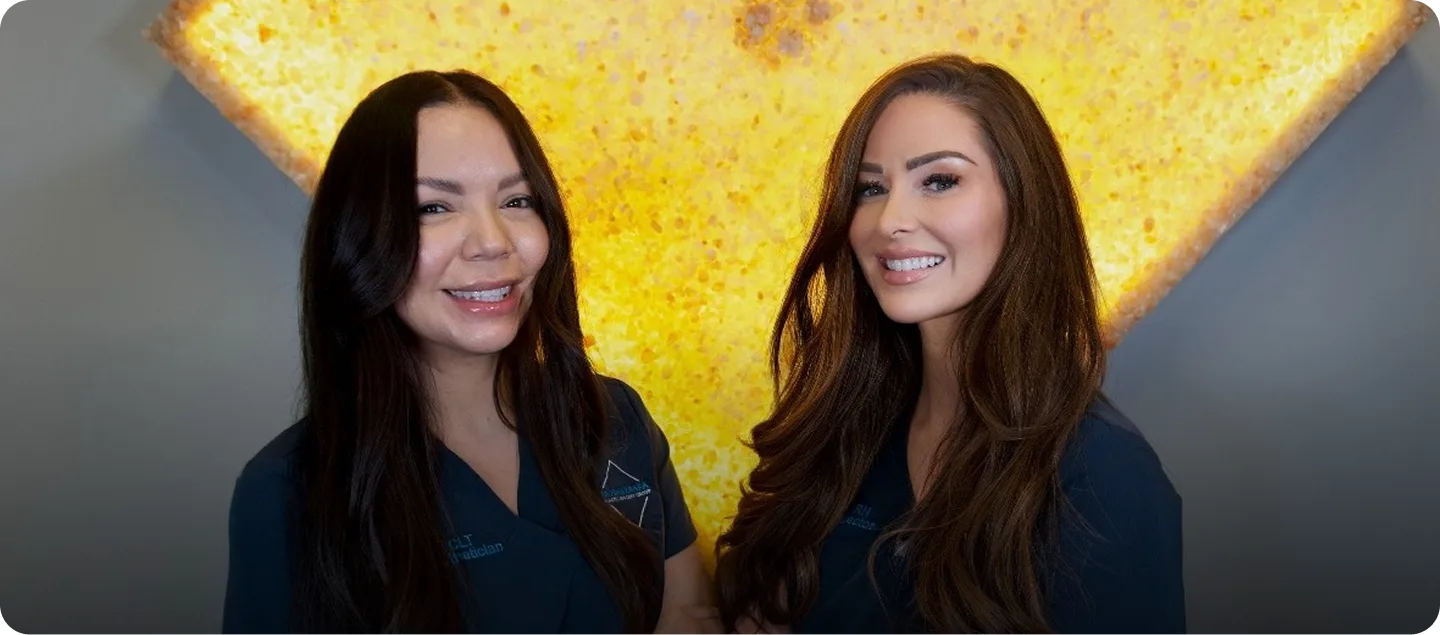 Two female staff members in dark blue scrubs smiling at camera against yellow textured background
