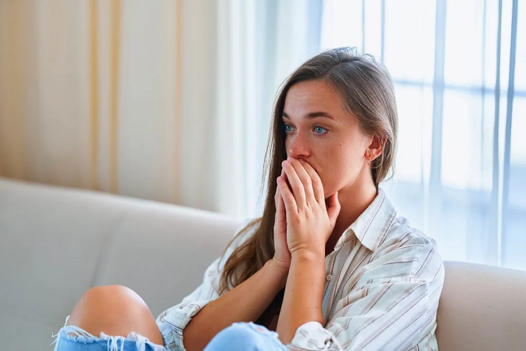 Woman sitting on a couch near a window, looking troubled with her hand near her face