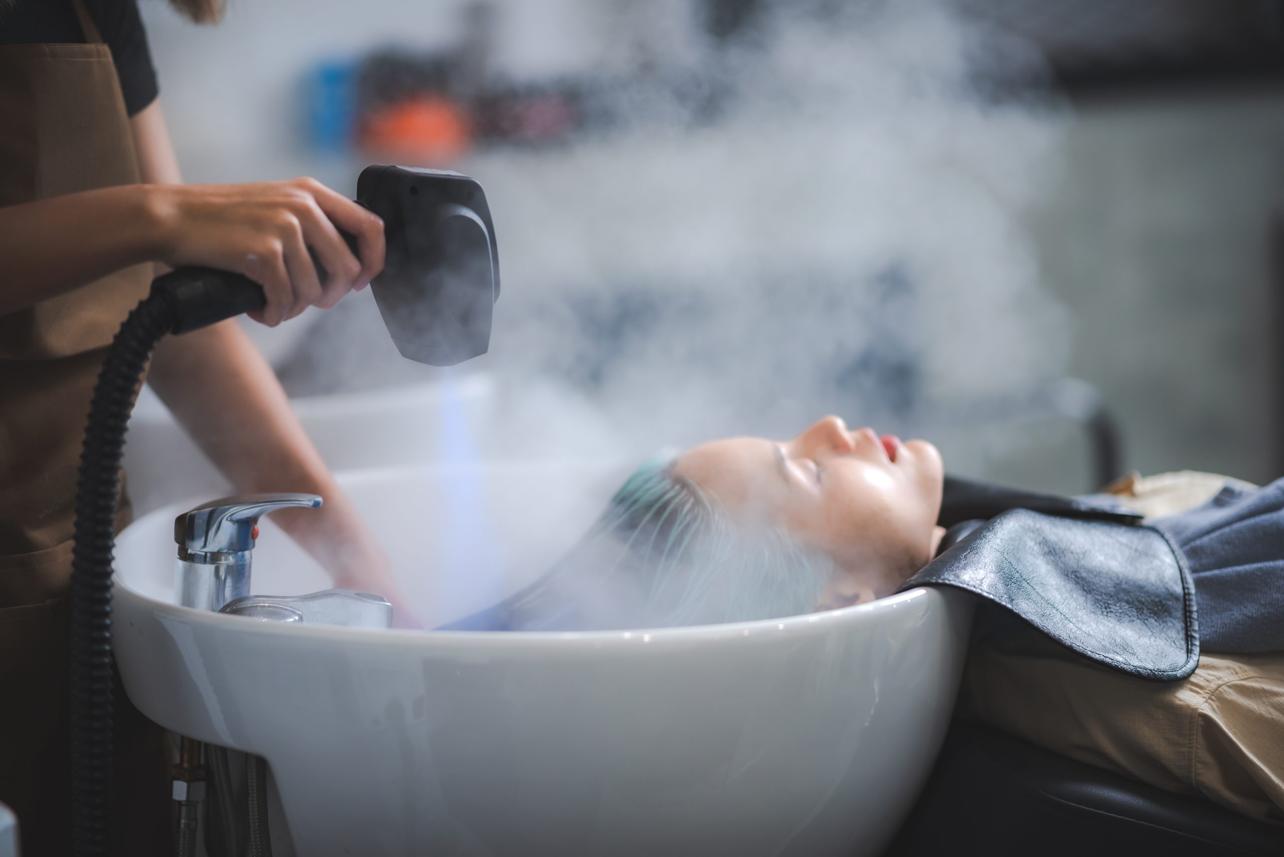 Stylist applying steam treatment to client's hair over white basin