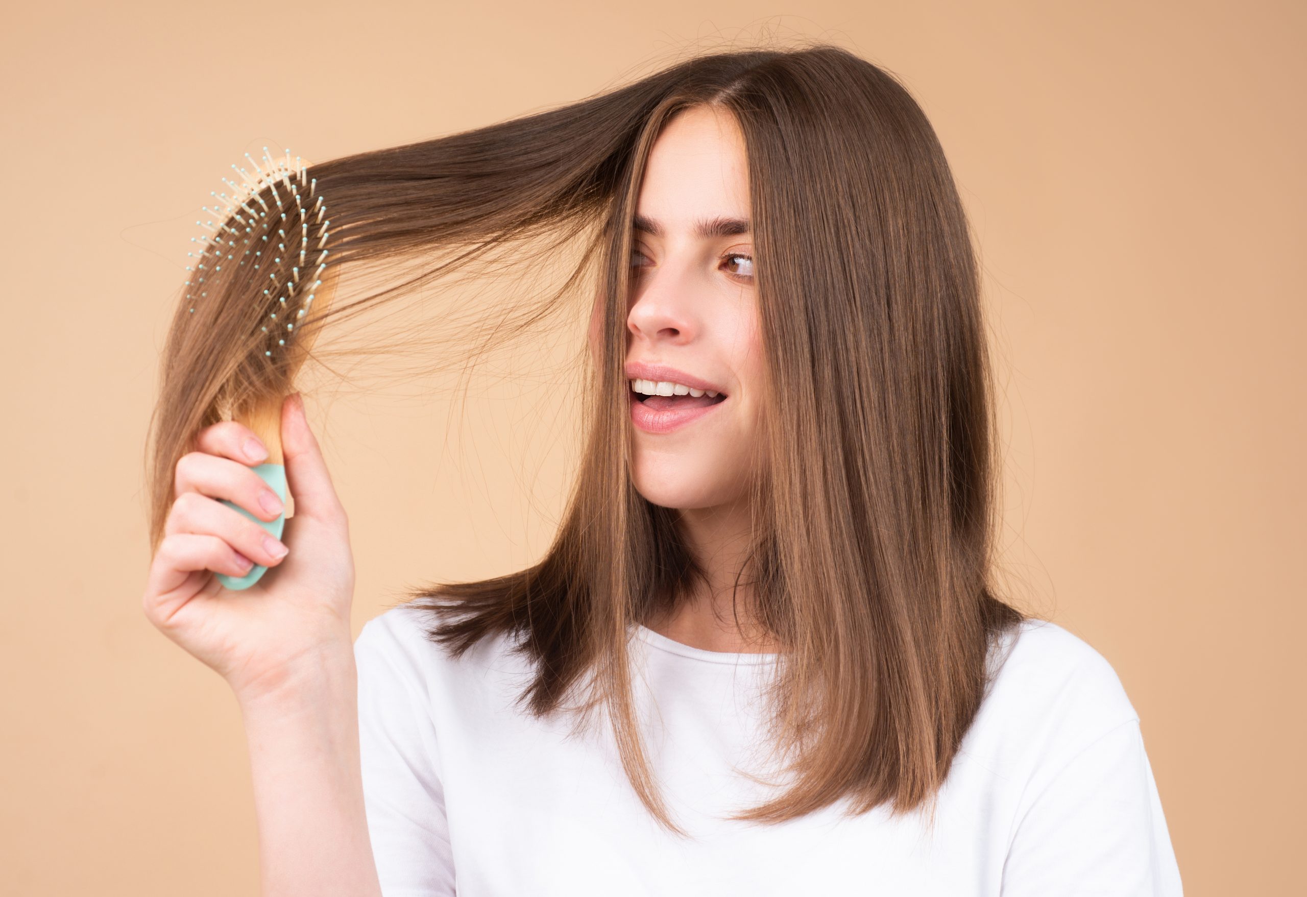 Woman brushing her long healthy brown hair with a paddle brush against a beige background