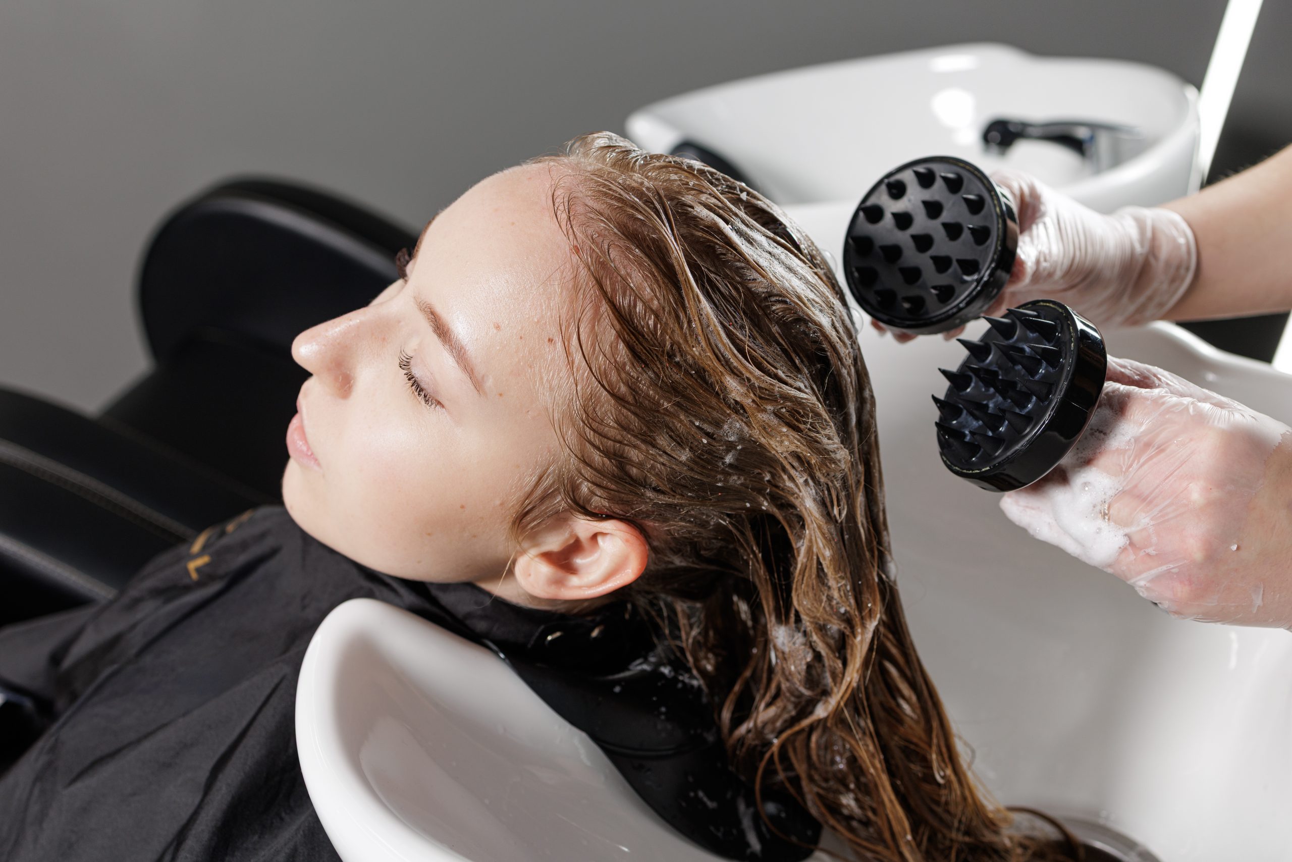 Stylist using black scalp massage tools on client's wet hair at salon basin