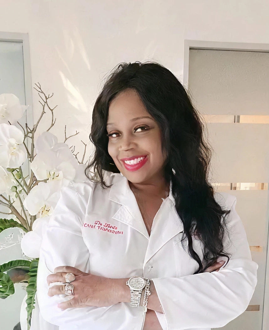 Woman in white lab coat smiling at camera with arms crossed next to white orchid flowers