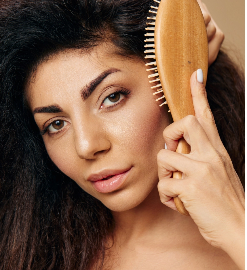 Woman brushing her long dark hair with wooden paddle brush, smiling at camera