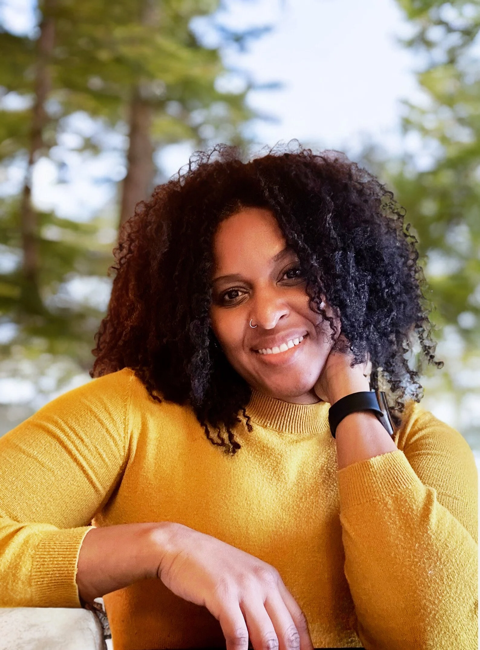 Woman smiling outdoors wearing yellow sweater with trees in soft-focus background