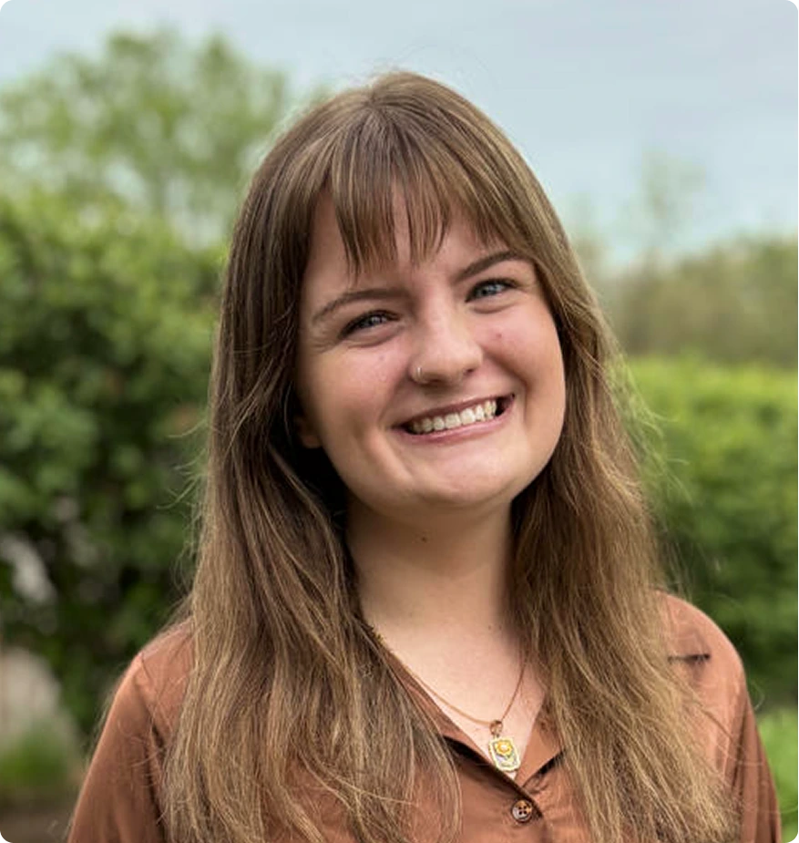 Therapist with brown hair and bangs smiling outdoors with green foliage background