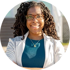 Woman wearing glasses and white coat over teal top, smiling at camera