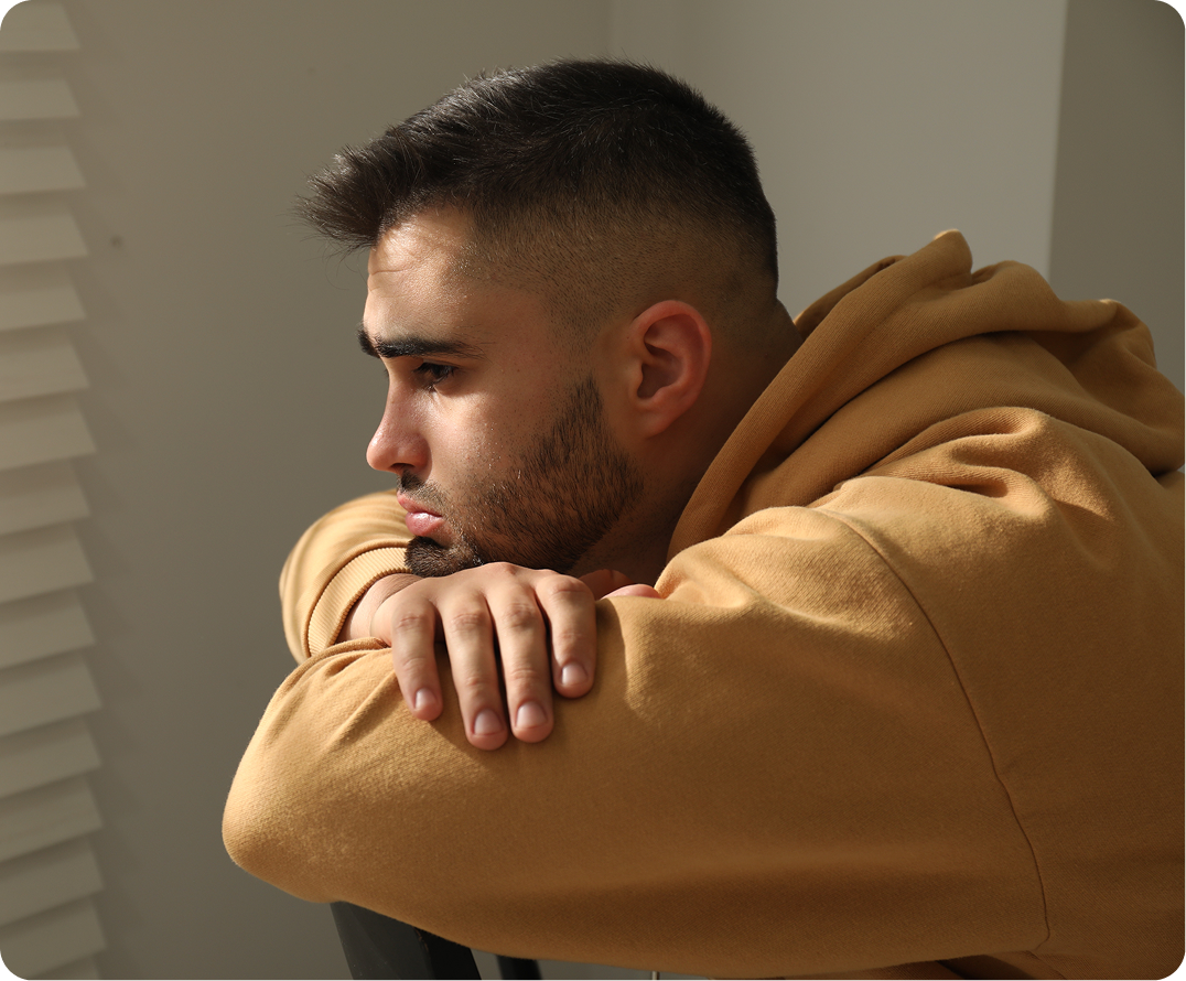 Man in tan hoodie hugging himself with downward gaze, appearing sad or withdrawn