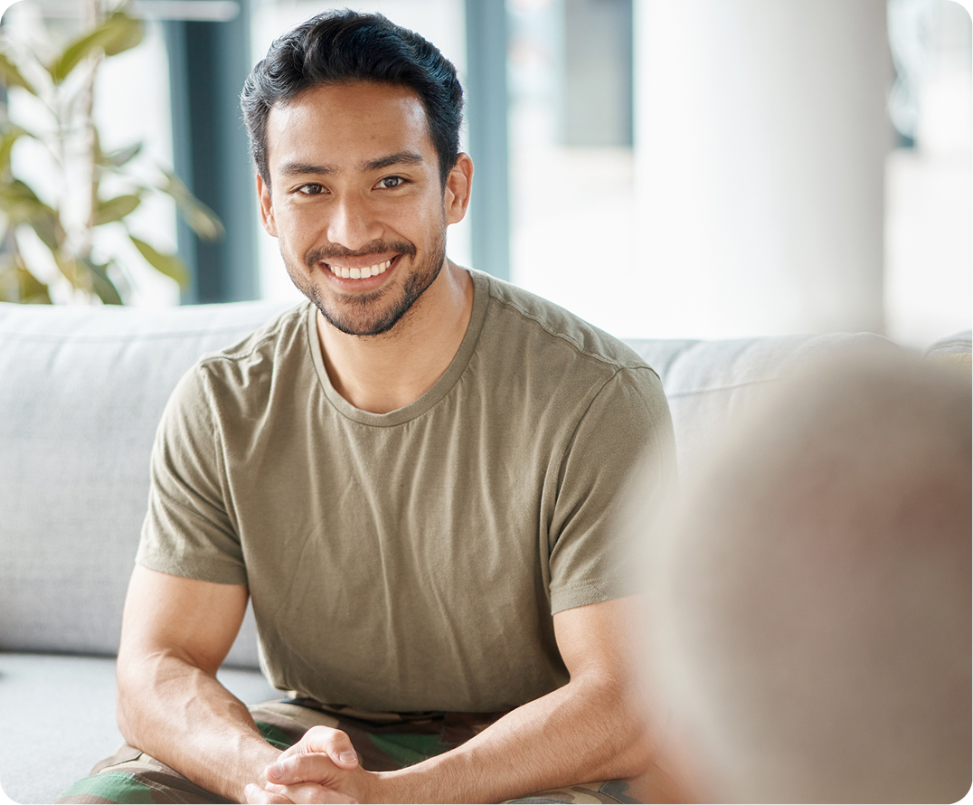 Smiling man in tan shirt sitting on white couch in bright room with plants