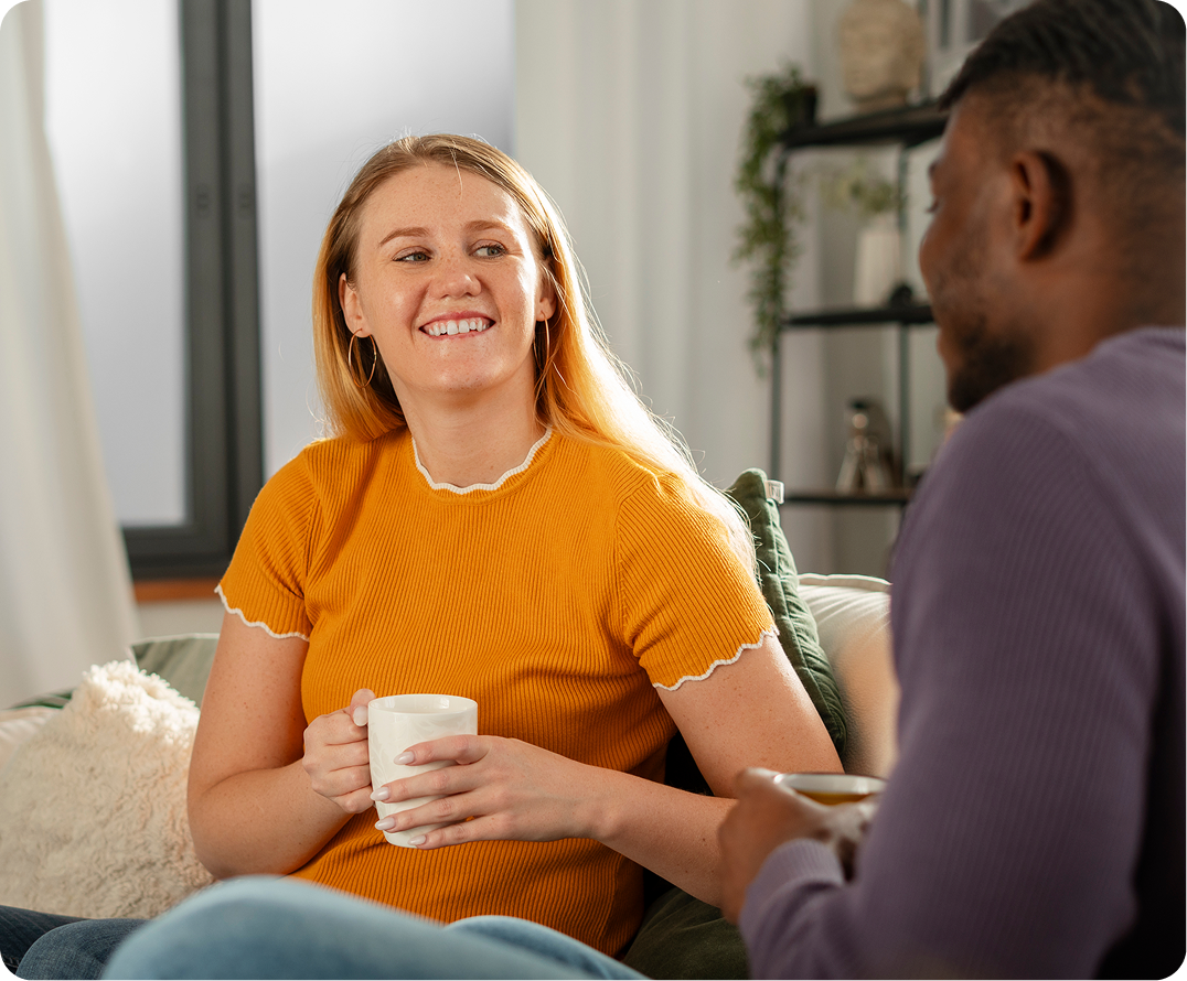 Woman in orange shirt holding cup smiling at man during conversation in bright living room