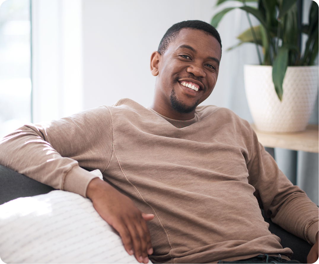 Man smiling while sitting on couch with potted plant on shelf behind him