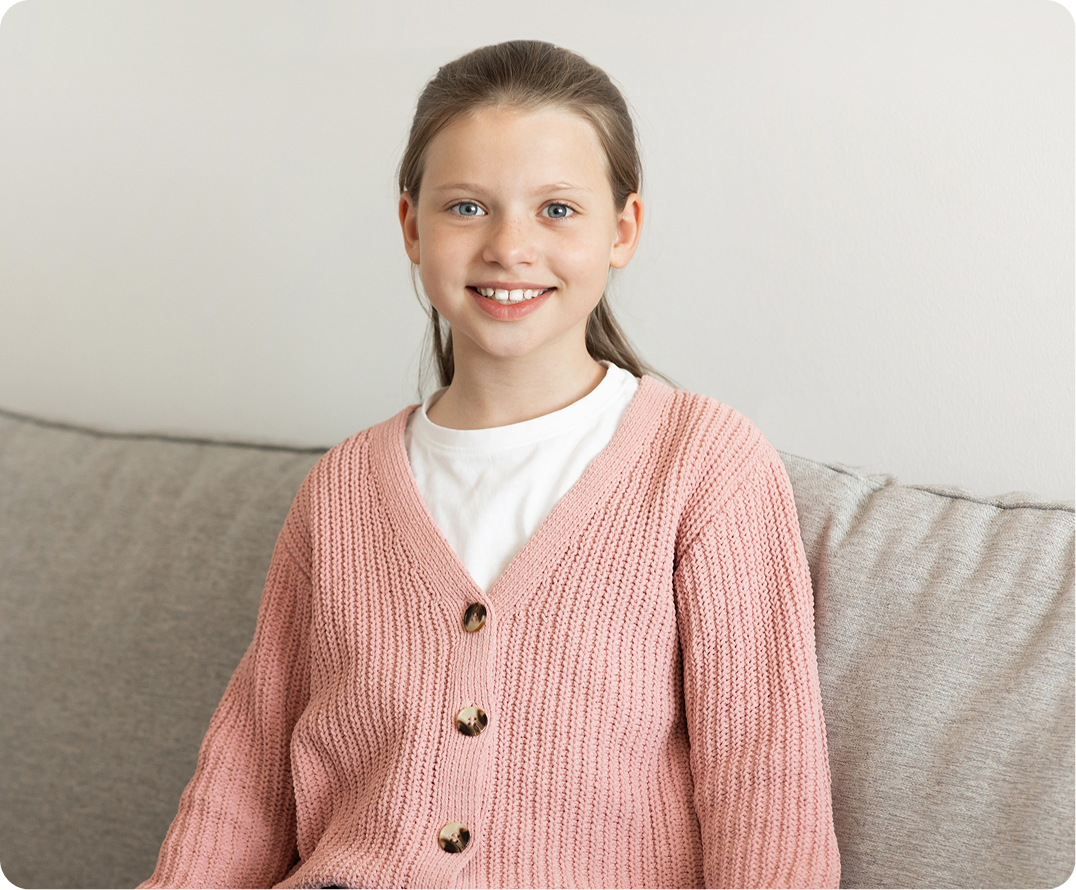 Girl sitting on couch wearing pink cardigan, smiling at camera