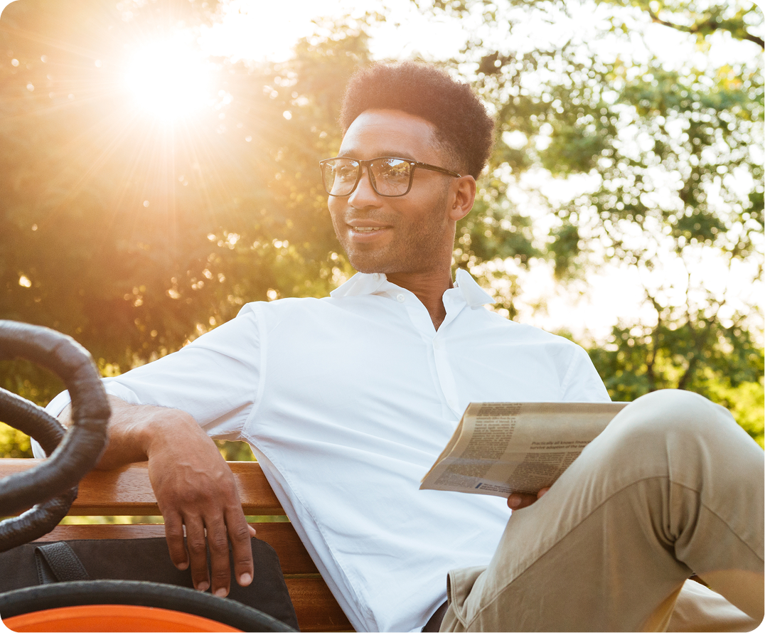 Man wearing glasses reading newspaper outdoors on bench in golden sunlight