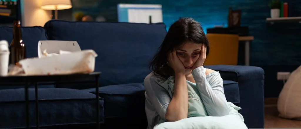 Woman sitting on couch with head in hand, looking downward with distressed expression in dimly lit living room