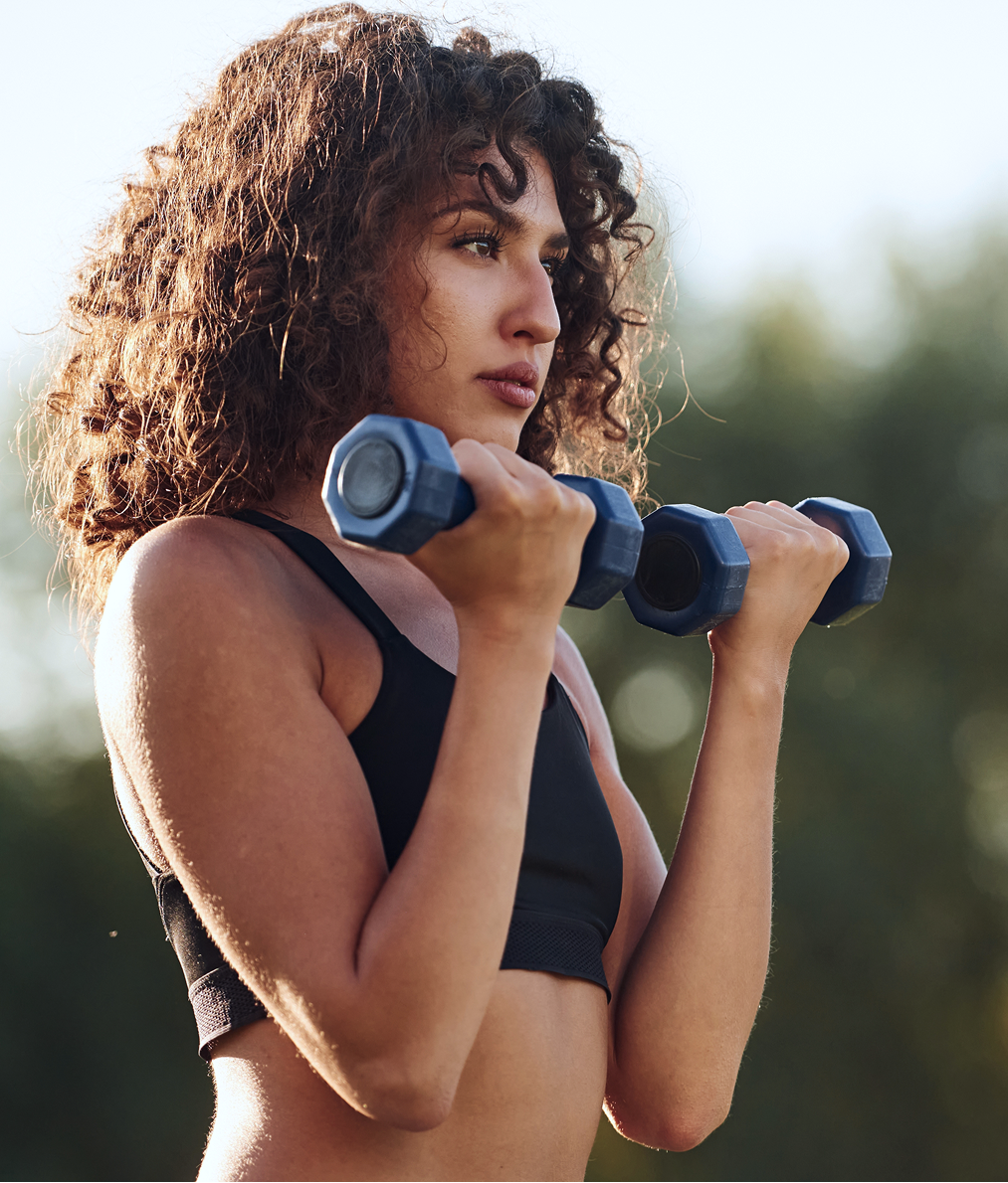 Woman with curly hair holding blue dumbbells in exercise stance outdoors