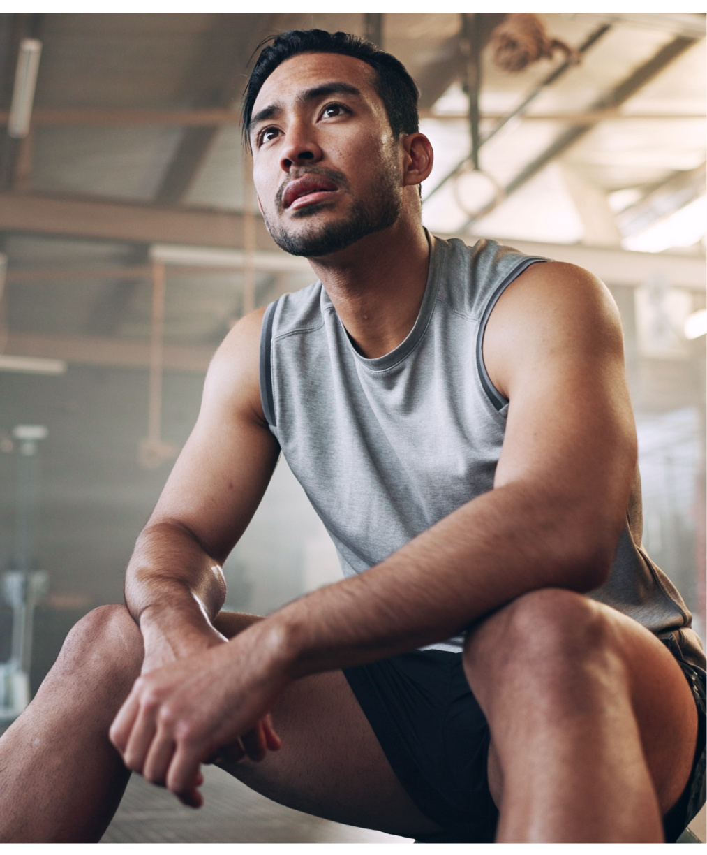 Man in gray sleeveless shirt sitting thoughtfully in bright modern gym setting