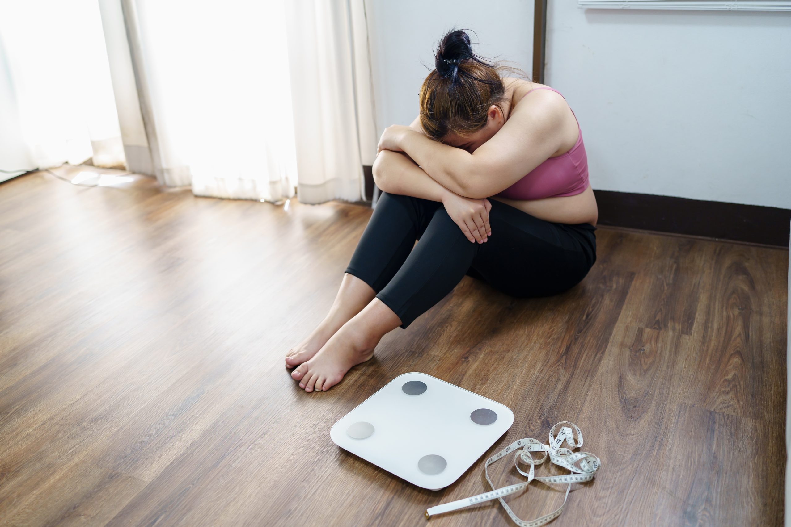 Woman sitting on floor with head down next to a scale and measuring tape, expressing frustration with weight loss