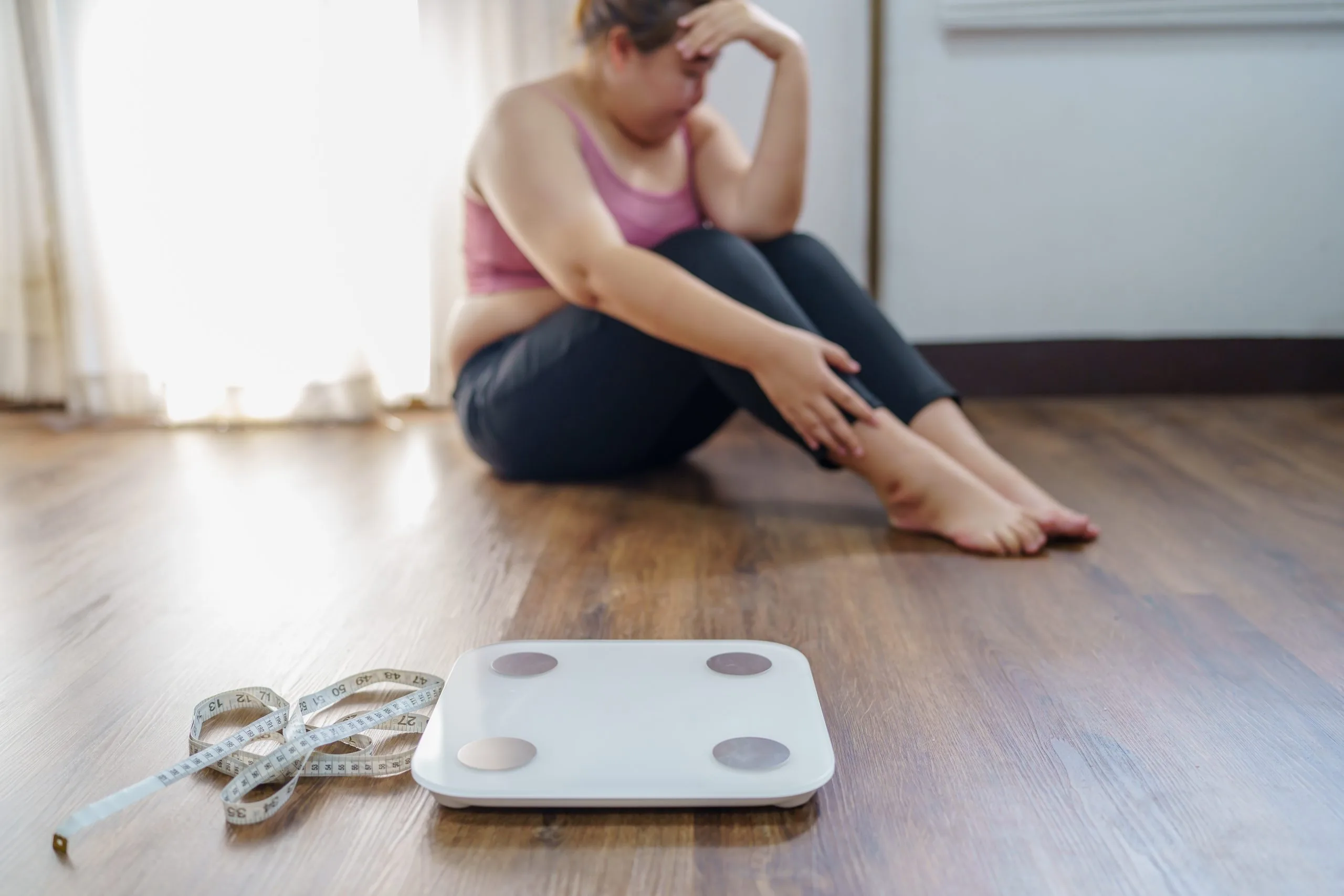 Woman sitting on floor looking discouraged with scale and measuring tape in foreground
