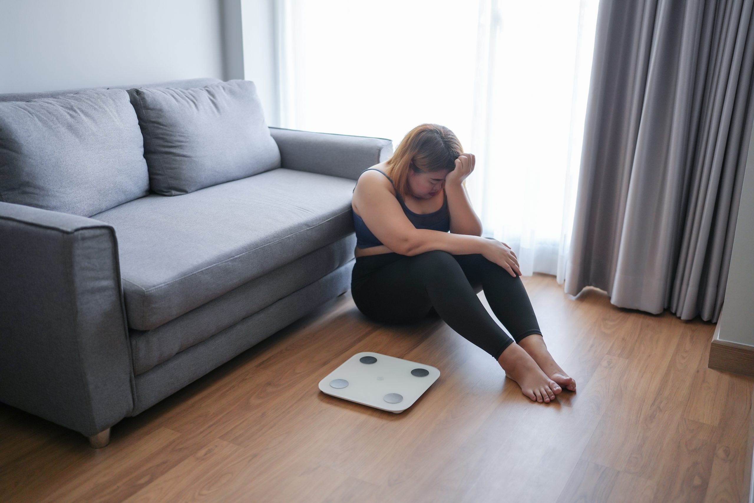 Woman sitting on floor next to scale, looking discouraged during weight loss journey