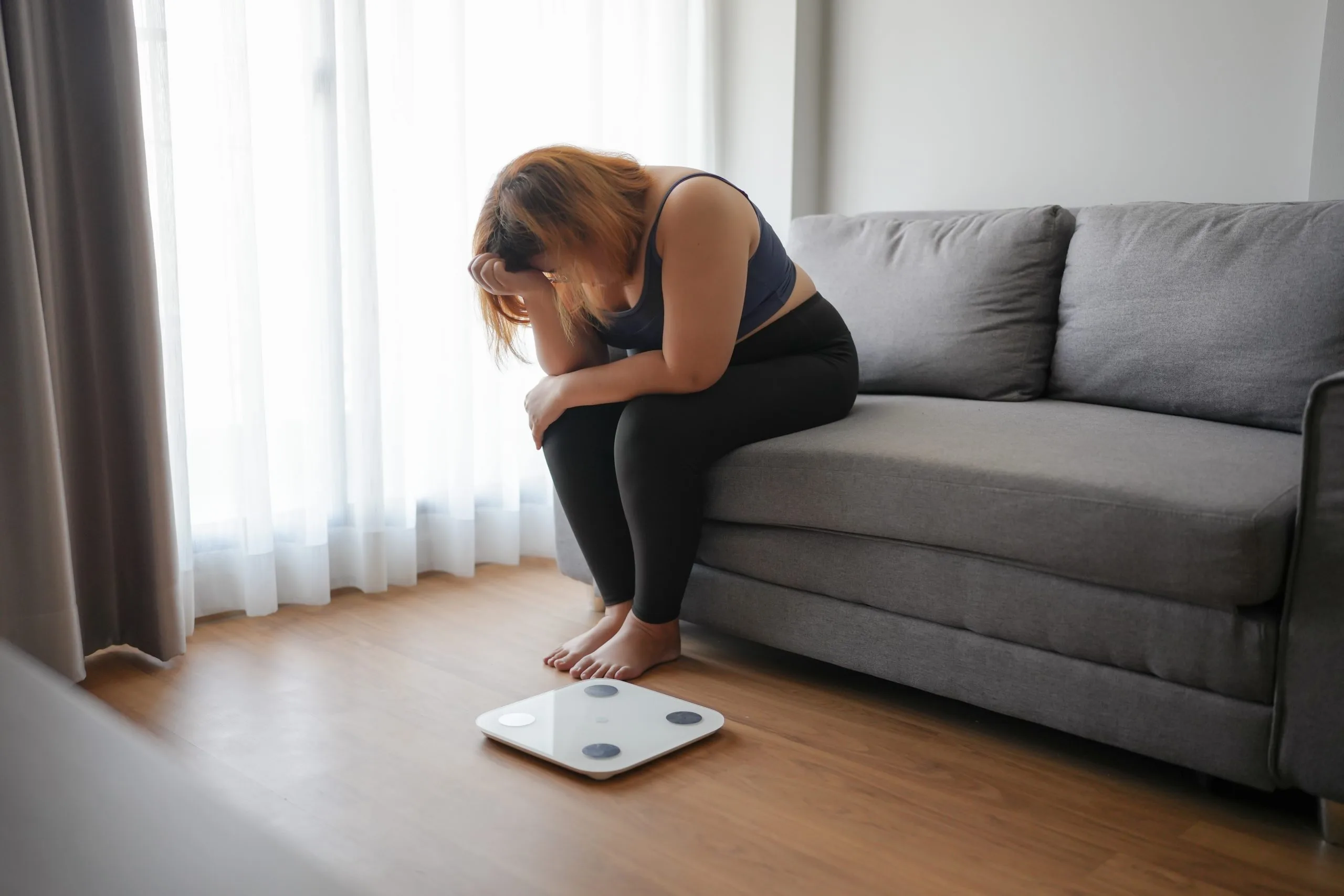 Woman sitting on couch looking down at a bathroom scale on the floor with an upset expression
