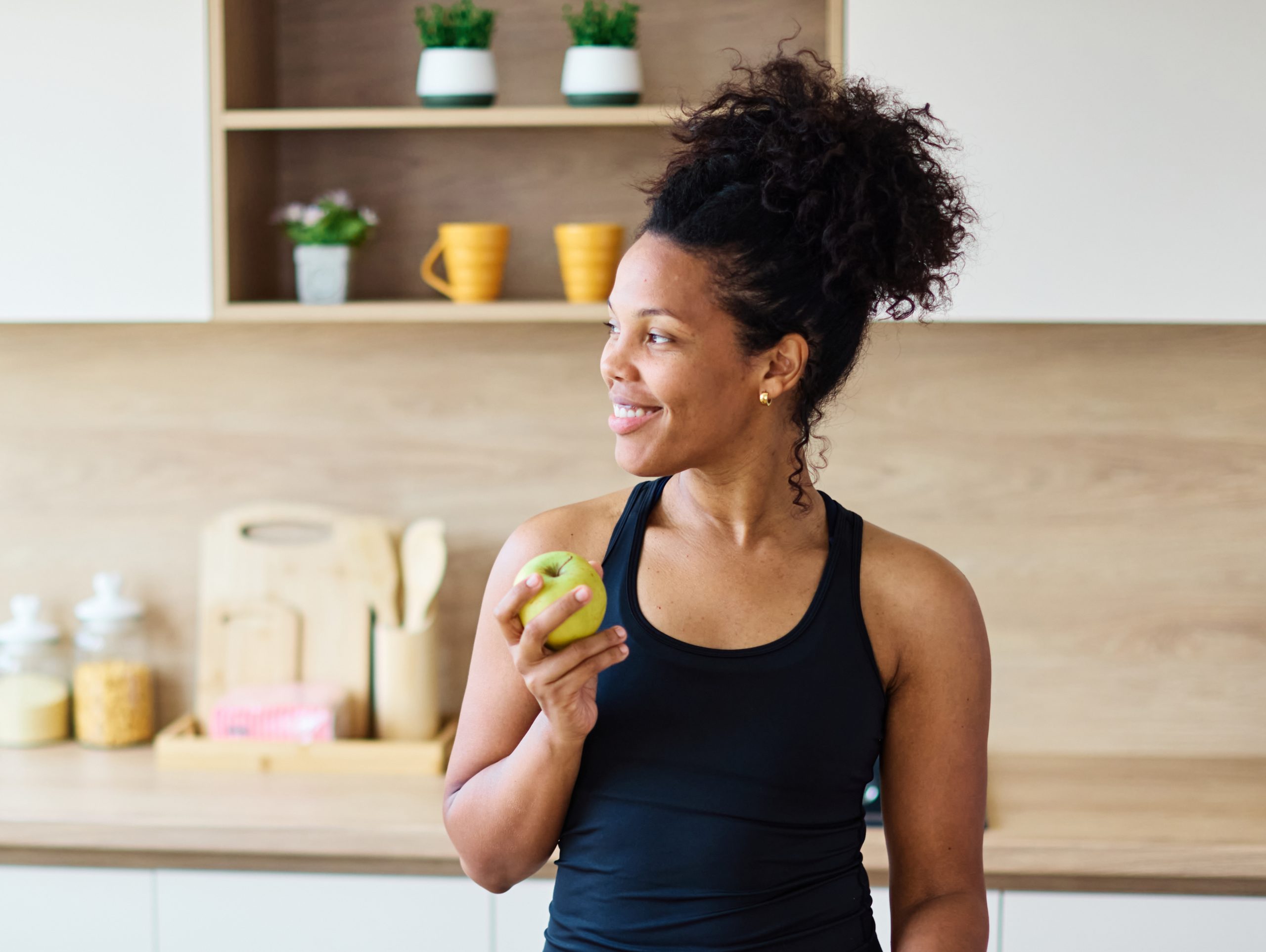Woman in black tank top holding a green apple in a modern kitchen with open shelving