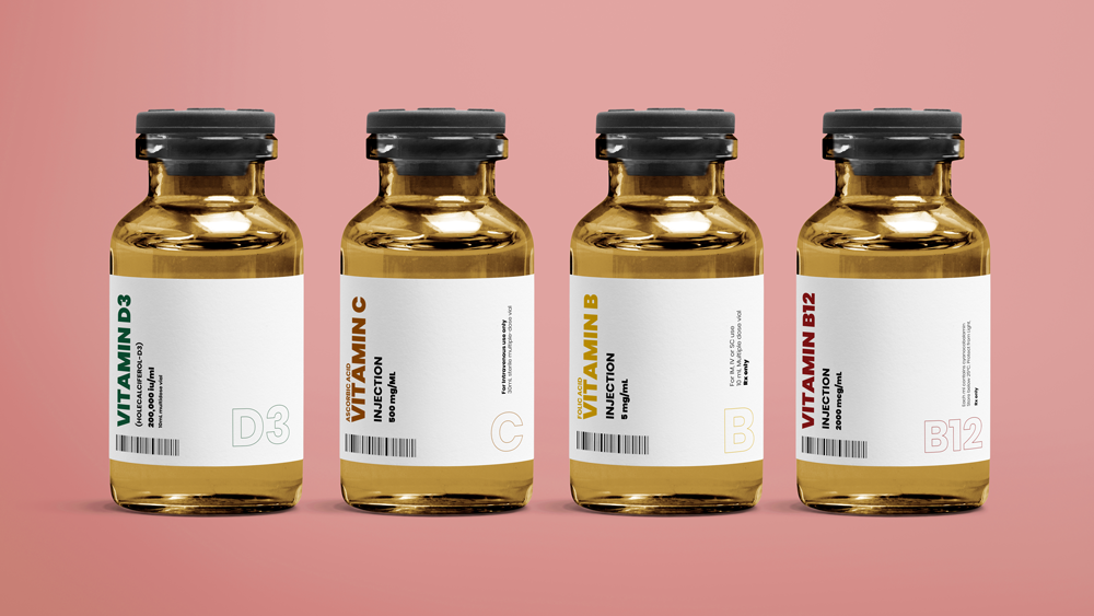 Four glass vials of golden liquid labeled Vitamin C, Vitamin D3, Vitamin B, and Vitamin B12 against a pink background