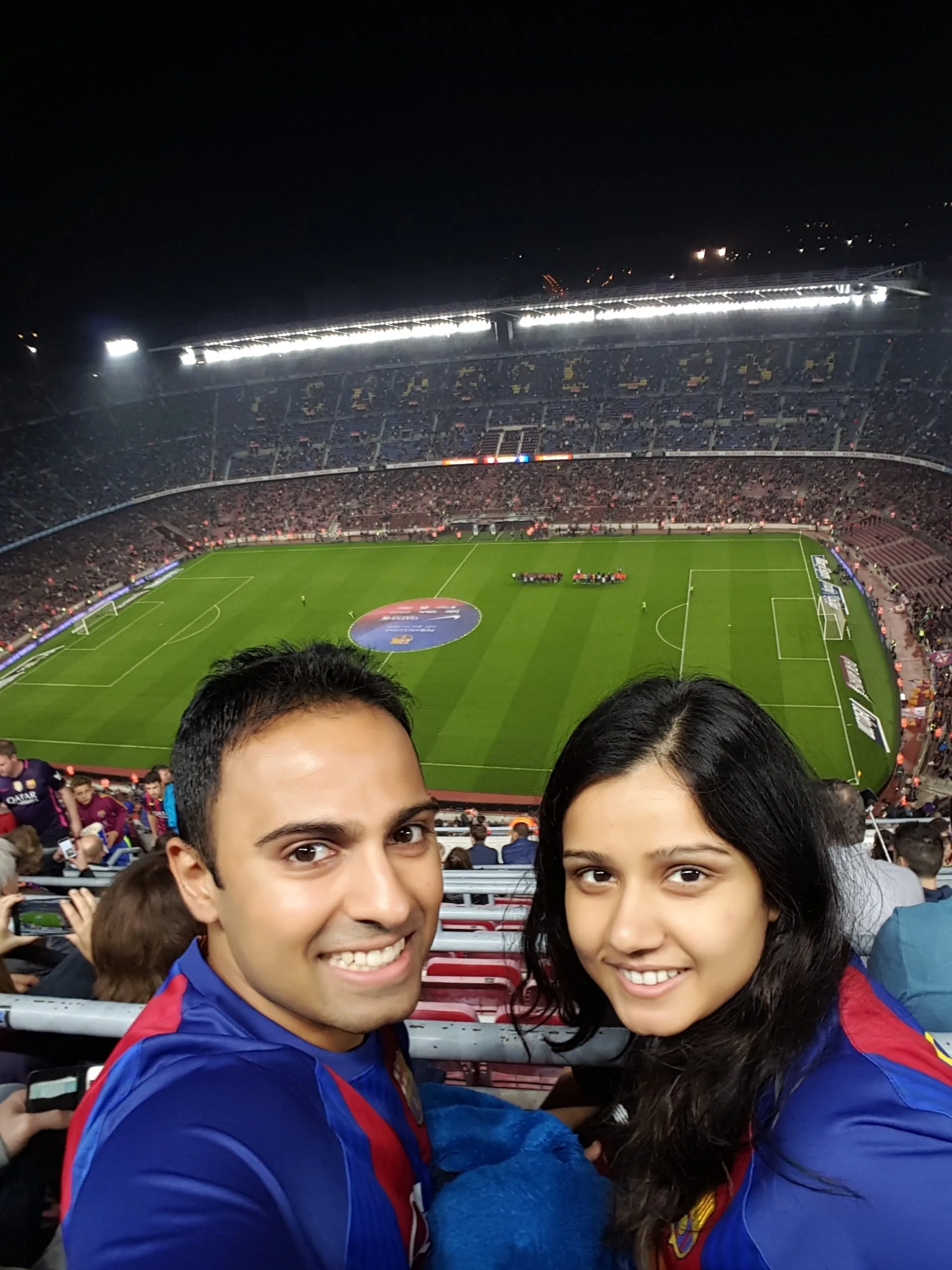 Two people in blue and red sports jerseys smiling at a crowded soccer stadium