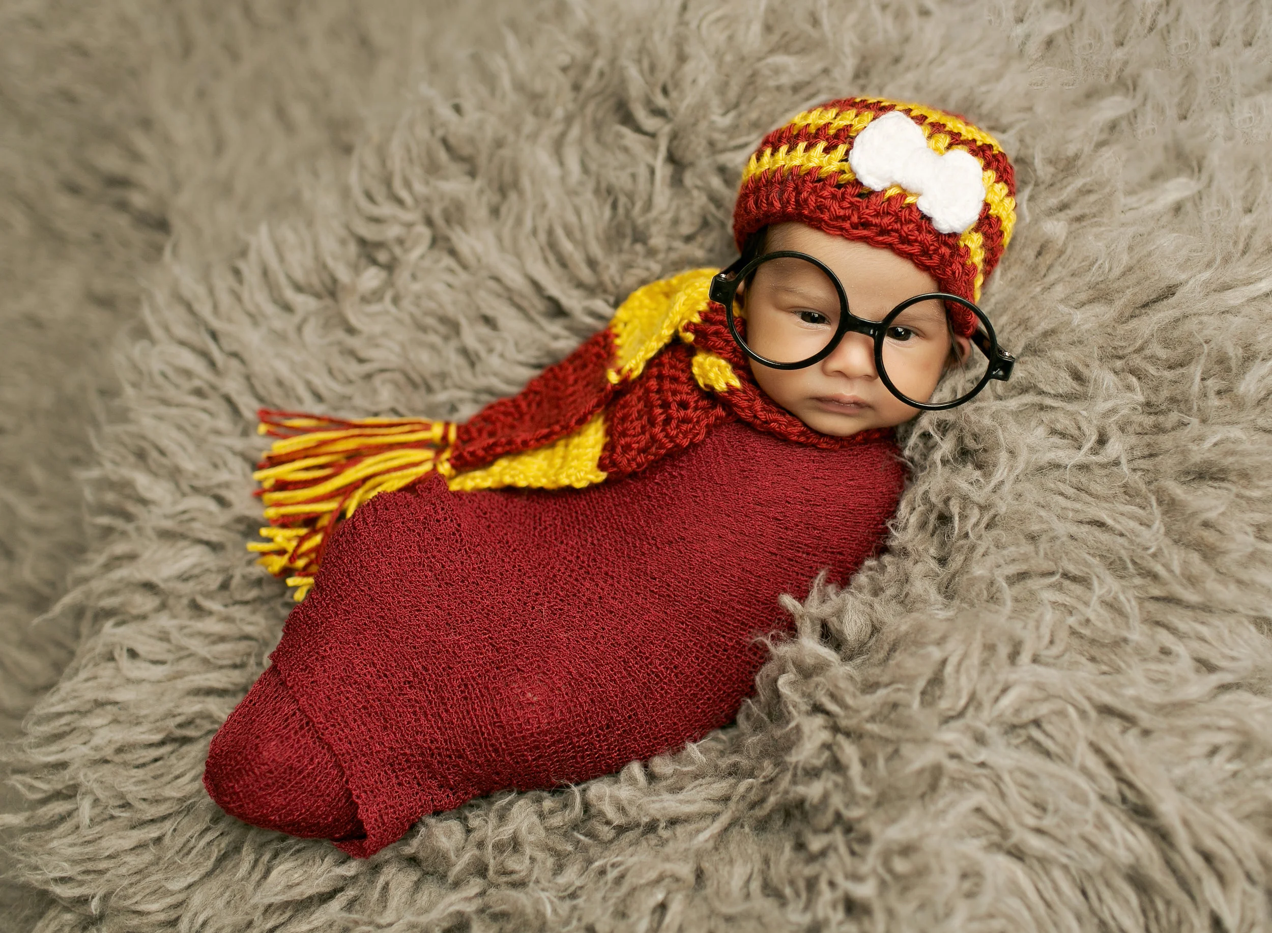 Newborn wearing oversized red and yellow knitted scarf and eyeglasses on gray textured blanket