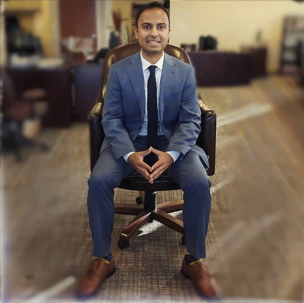 Man in blue suit and tie seated in office chair with blurred office background
