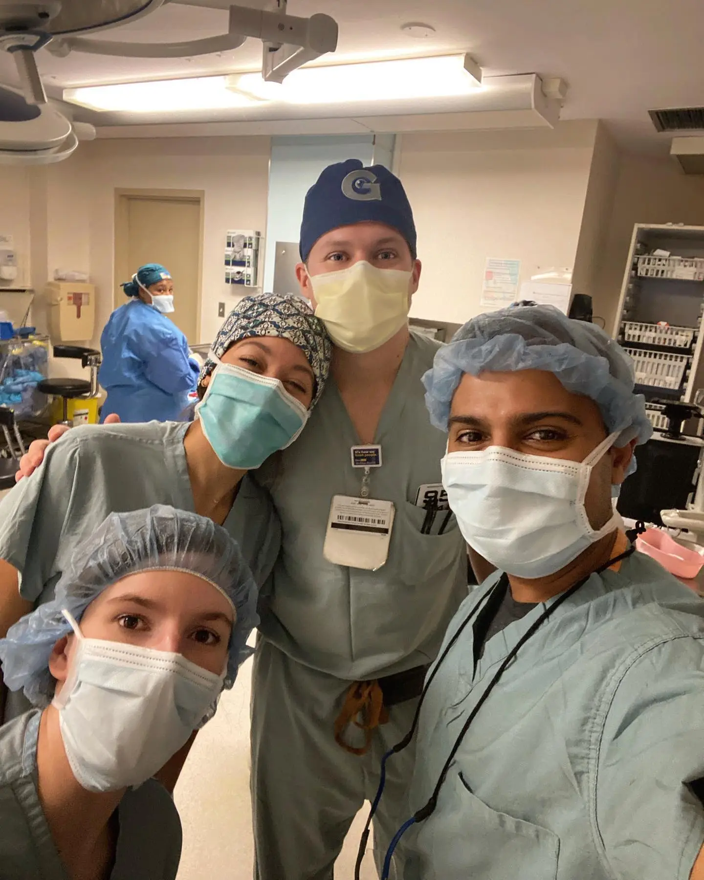 Four surgical team members wearing masks and head coverings posing together in operating room