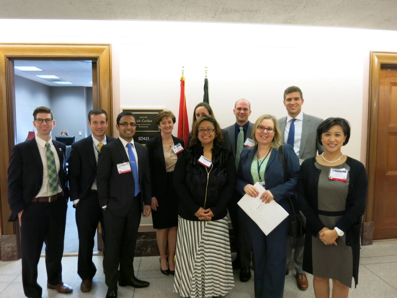 Nine professionals in business attire at an indoor event with name badges and flags