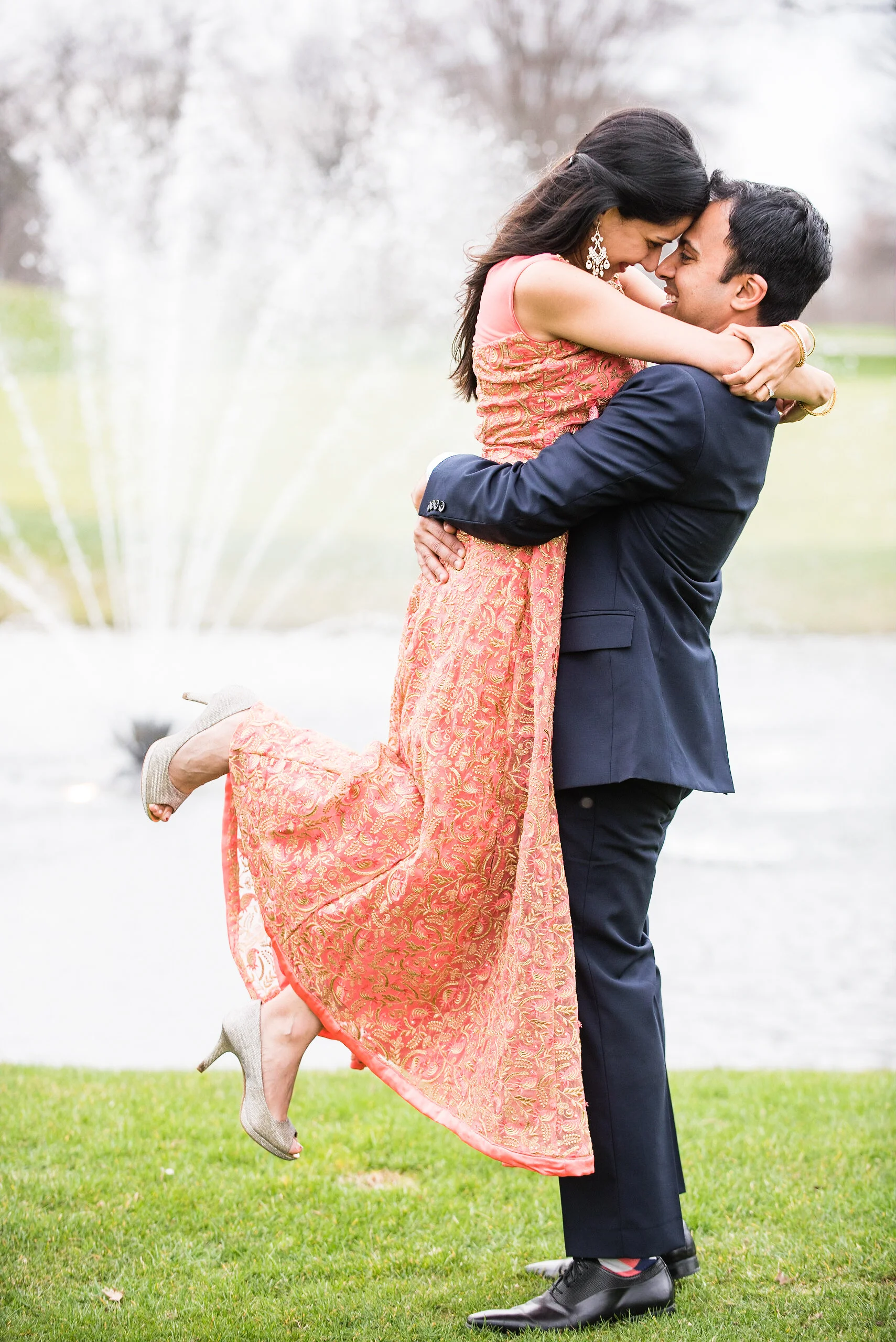 Man lifting woman in coral patterned dress outdoors with blooming white trees in background